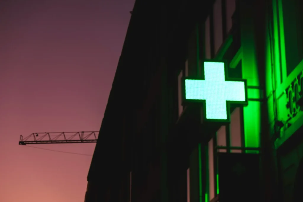 Grün leuchtendes Apothekenkreuz an einer Hauswand in der Abenddämmerung, links ein Baukran vor rosa Himmel
